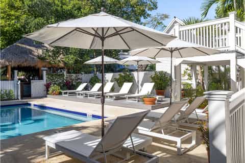 Resort pool with thatched cabana, white lounge chairs under large umbrellas, and charming coastal architecture.