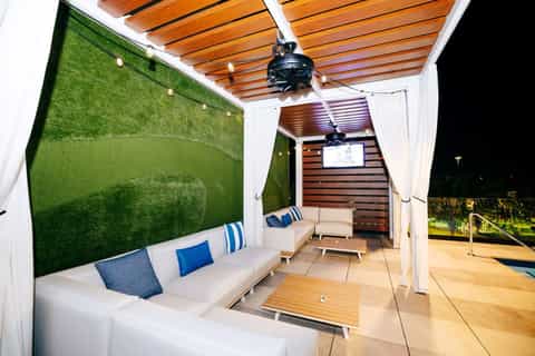 Covered outdoor lounge area with white furniture, green wall feature, and wooden ceiling