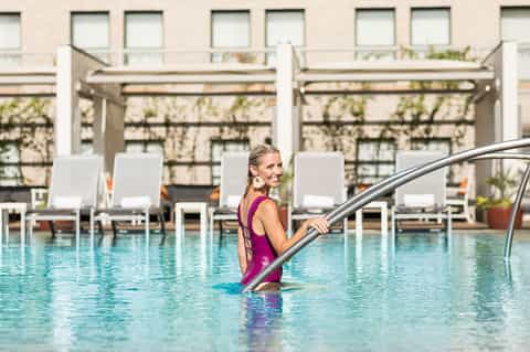 Woman in purple swimsuit entering an outdoor pool with white lounge chairs and pergolas in background