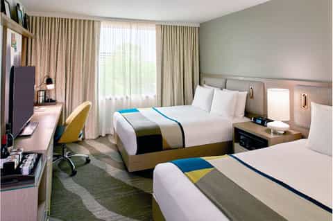 Hotel room with two queen beds, blue and yellow accents, work desk, and floor-to-ceiling curtains