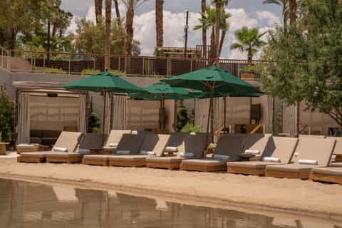 Beachside cabanas with green umbrellas, daybed loungers, curtains, and palm trees along sandy shore