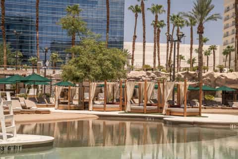 Resort pool area with cabanas, palm trees, lounge chairs, and modern high-rise buildings nearby