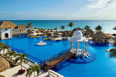 Large lagoon-style resort pool with white gazebo, thatched-roof palapa structures, and ocean views