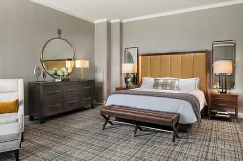 Contemporary hotel bedroom with tan upholstered headboard, dark dresser, round mirror, and neutral décor