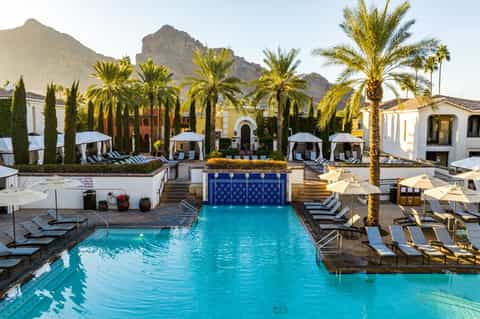 Resort pool courtyard with turquoise water, palm trees, white umbrellas, and mountain backdrop