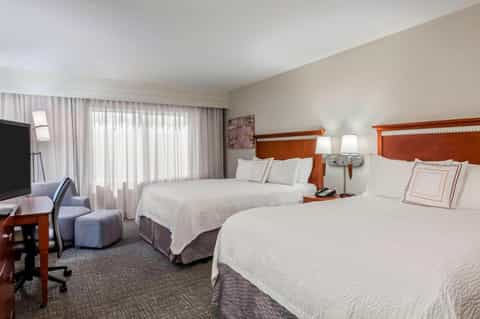 Hotel room with two queen beds, orange headboards, desk area, and sheer curtain-lined windows