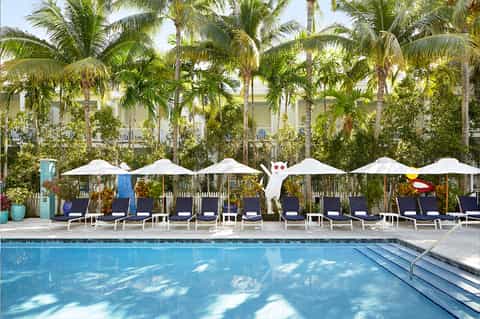 Resort pool with white umbrellas, lounge chairs, palm trees, and colonial-style building in background