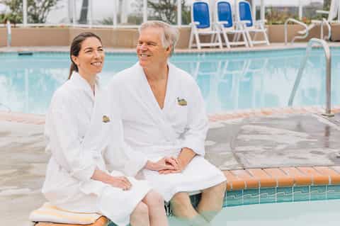 Couple in white robes sitting poolside at resort with lounge chairs and water features visible