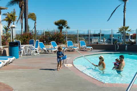 Rooftop pool deck with families swimming, lounge chairs, and ocean views behind glass railing