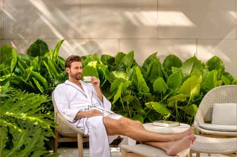 Man in white spa robe sitting outdoors near large green tropical plants on terrace