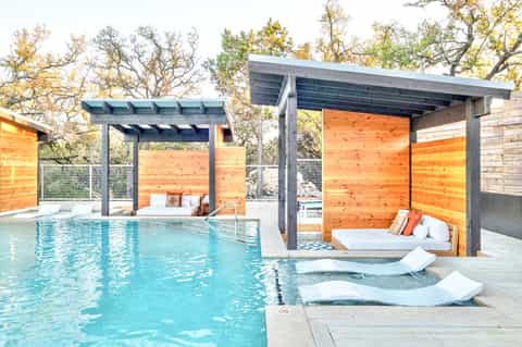 Contemporary pool with wooden cabanas, concrete pergolas, and white lounge chairs
