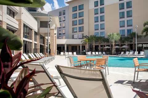 Resort pool deck with lounge chairs, palm trees, and multi-story hotel buildings in sunny weather