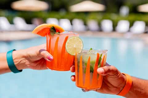 Two colorful tropical cocktails with citrus garnishes by pool with loungers and umbrellas in background