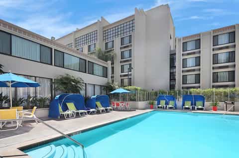 Modern hotel pool with blue canopy loungers, umbrellas, and multi-story building backdrop