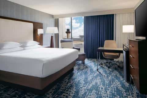Modern hotel bedroom with king bed, dark wood furniture, blue curtains, desk area, and city views