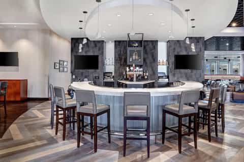 Modern hotel bar with curved white counter, dark gray tile wall, pendant lighting, and bar seating