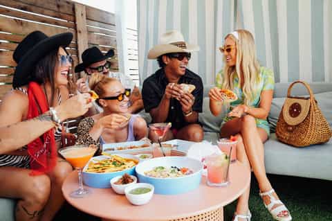 Group of friends relaxing on outdoor lounge with food, drinks, and cocktails in casual beachy setting