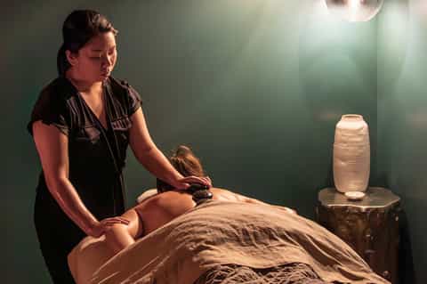 Spa therapist performing hot stone massage on client in peaceful green-walled treatment room with ambient lighting