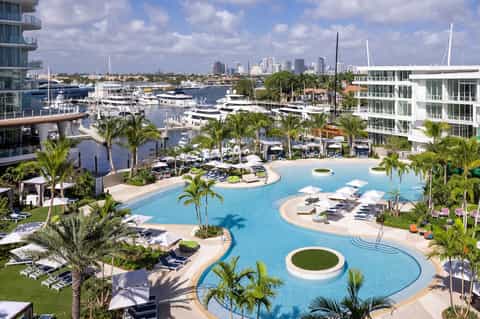 Waterfront resort pool with curved blue water, palm trees, loungers, marina with boats, and skyline in background