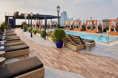 Rooftop pool deck with lounge chairs, wooden cabanas, pergola, and city skyline views