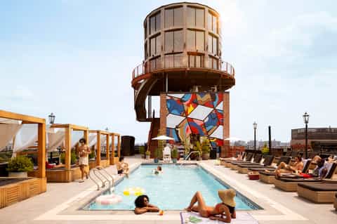 Rooftop pool with water tower sculpture, loungers, cabanas, and umbrellas overlooking city skyline