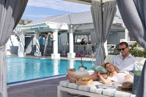 Couple relaxing on poolside cabana bed overlooking resort pool and Shores Bar facility