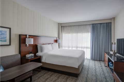 Hotel bedroom with queen bed, wood headboard, modern furnishings, and ocean-view balcony