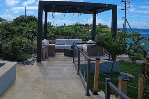 Oceanside terrace with black pergola, wicker seating, hanging lights, and tropical coastal views