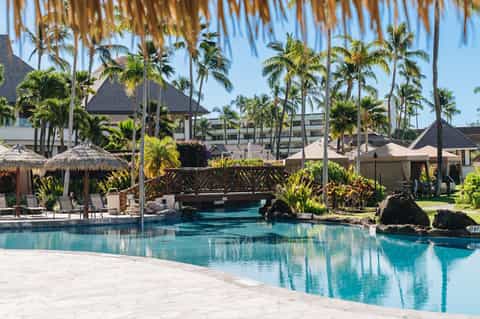 Resort pool with wooden bridge, palm trees, and tiki umbrellas under blue sky