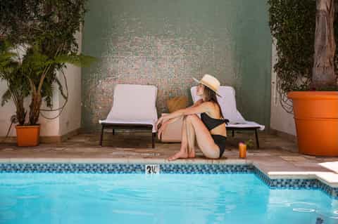 Woman relaxing on lounge chairs by turquoise pool with mosaic tile wall and potted plants