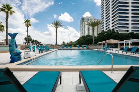 Resort pool with turquoise water, blue lounge chairs, palm trees, and tall buildings