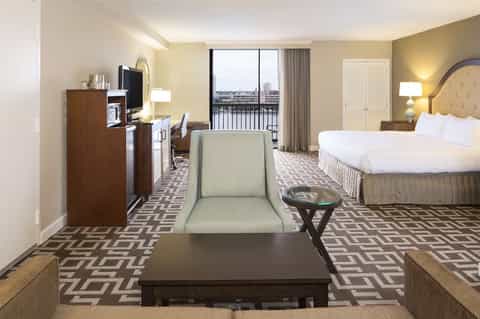 Spacious hotel room with queen bed, desk area, and waterfront balcony view with patterned carpet