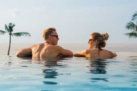 Couple relaxing in infinity pool overlooking ocean with palm trees on horizon