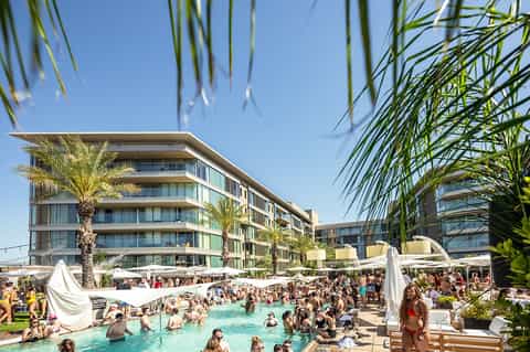 Resort pool deck with modern architecture, palm trees, white umbrellas, and crowds of guests swimming