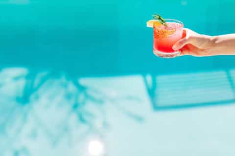 Hand holding a colorful cocktail with garnish by a bright turquoise pool