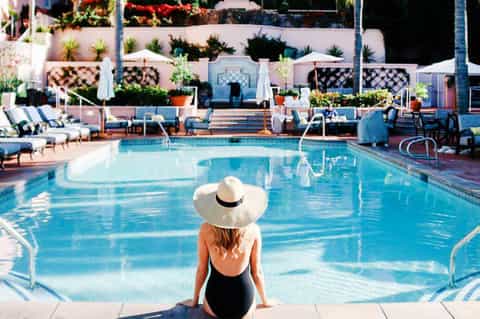 Woman in swimsuit and hat sitting poolside overlooking resort pool with pink buildings and tropical plants