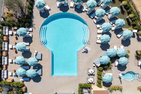 Aerial view of resort pool deck with kidney-shaped pool, blue umbrellas, lounge chairs, and palm trees