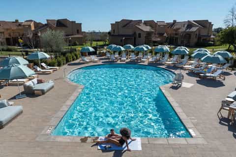Curved resort pool surrounded by blue umbrellas, lounge chairs, and Mediterranean-style villa buildings