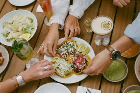 Overhead view of diners sharing Mexican food with cocktails at wooden table