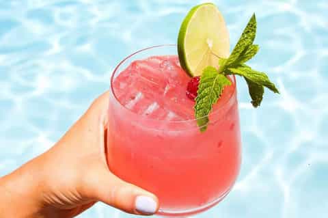 Tropical cocktail with coral-colored liquid, lime slice, and mint garnish held over pool water
