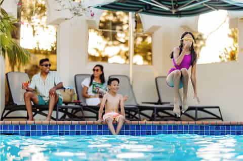 Family enjoying resort swimming pool with girl jumping in, adults relaxing poolside under umbrellas