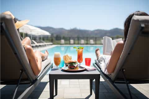 Couple relaxing poolside with cocktails, food, mountains visible in background