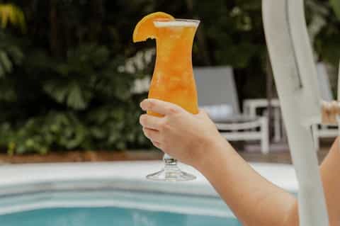 Hand holding tropical orange cocktail with orange slice garnish by resort pool