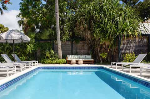 Resort swimming pool with blue water, white loungers, palm trees, wooden fence, and poolside seating