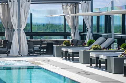 Modern rooftop lounge with gray loungers, white curtains, glass railings, and panoramic forest and sky views