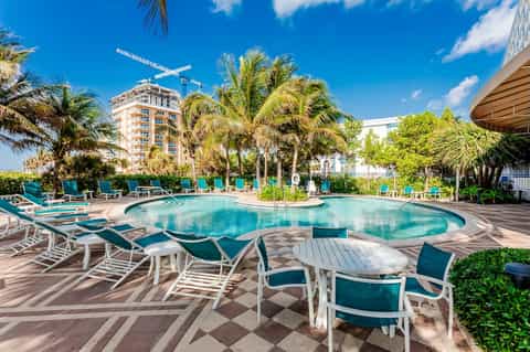 Resort pool area with turquoise water, palm trees, white and teal lounge chairs, and high-rise building