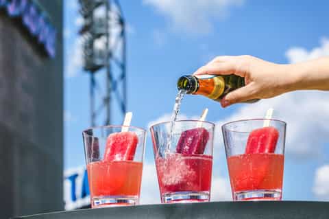 Hand pouring sparkling wine into three glasses with red popsicles against blue sky and city buildings