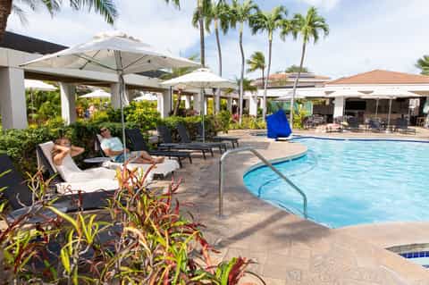 Resort poolside with lounge chairs under umbrellas, palm trees, and tropical landscaping on sunny day