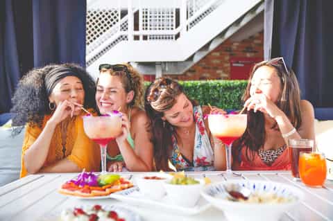 Four women enjoying colorful cocktails and appetizers at an outdoor poolside gathering