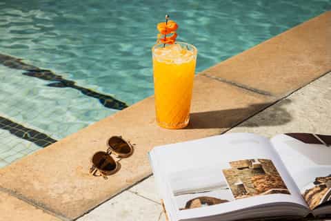 Poolside refreshment with yellow cocktail, sunglasses, and travel magazine on concrete deck